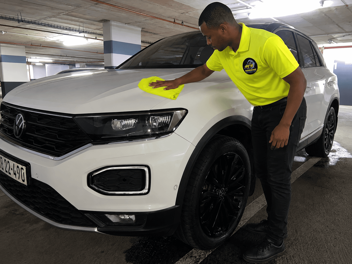 Man in yellow shirt wipes the hood of a white SUV with a yellow cloth.