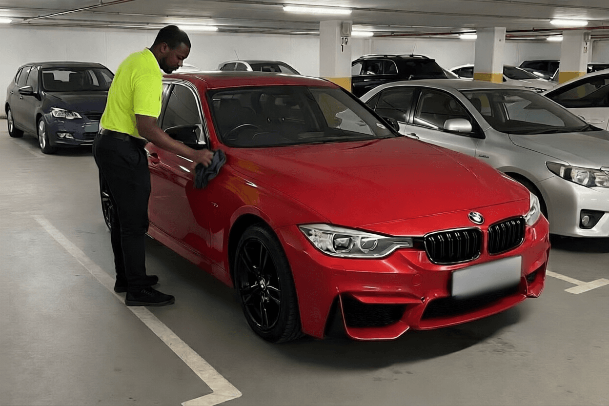 Man in neon yellow shirt cleaning a red BMW sedan in a parking garage.